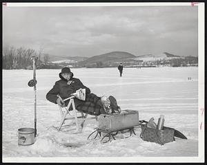Last Word in fishing comfort is used by Colvin Sykes as he fishes through the ice on Pontoosuc Lake, Pittsfield. He is equipped with a folding chair, a picnic lunch basket, his tackle box for a foot rest and some reading material. All he needs is some cooperation from the fish.