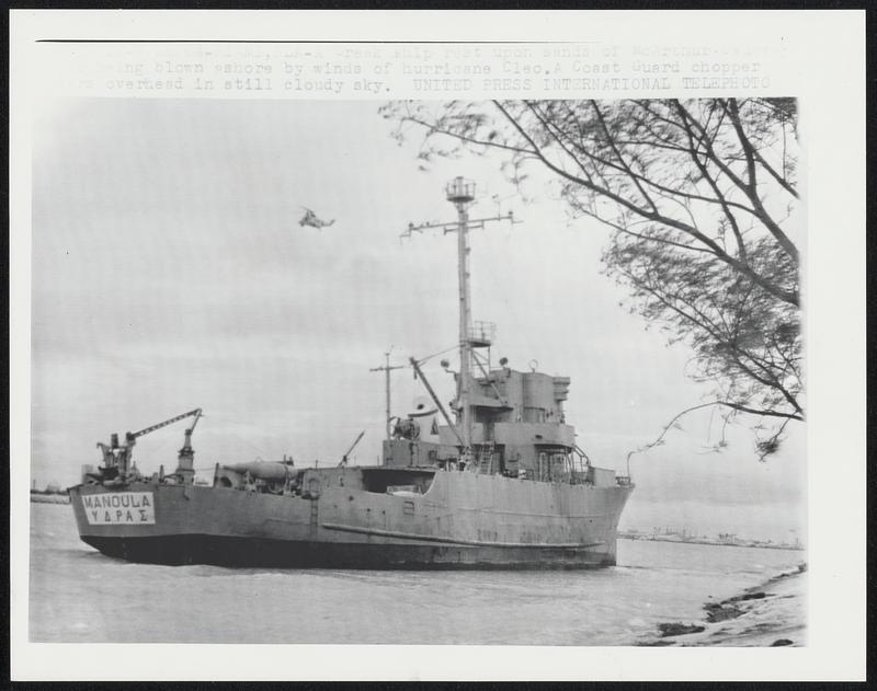 A Greek ship rest upon sands of McArthur Causeway being blown ashore by winds of hurricane Cleo. A Coast Guard chopper hovers overhead in still cloudy sky.