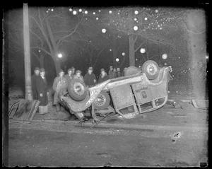 People stand behind an overturned automobile with "Longwood 5300" on its side
