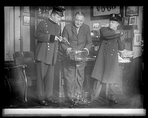 A man in police uniform pointing a gun at a bowl while another man in police uniform holds himself back