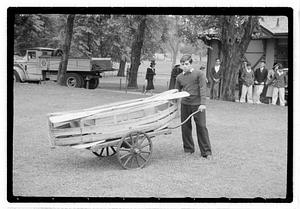 A young man pushing a cart with a wooden boat and oars on top