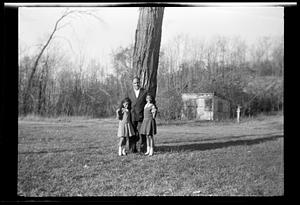 A man and two girls stand under a tree, shed in background