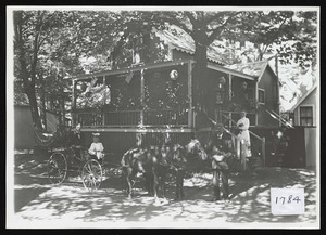 Two seater wagon drawn by two horses carrying man and girl, also man and woman with cat in front of victorian cottage named Mapleshade and decorated with flags and lanterns