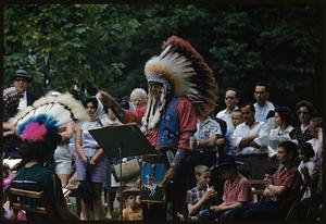 Man in feathered headdress conducting music while crowd watches