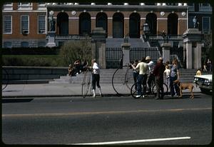 A group of people, two with penny-farthings and one with a normal bicycle in front of the Massachusetts State House