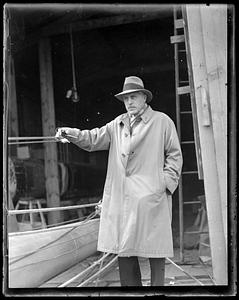 A man in a trench coat and hat stands holding a pipe