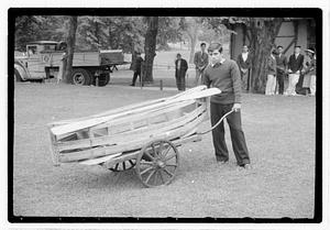 A young man pushing a cart with a wooden boat and oars on top