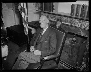 Gov. Christian Herter seated next to flag in front of fireplace