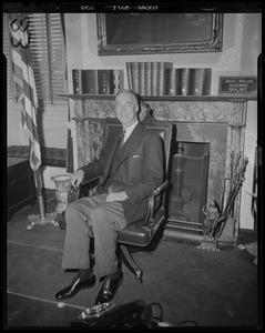 Gov. Christian Herter seated next to flag in front of fireplace