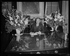 Gov. Christian Herter seated behind desk with large floral arrangements