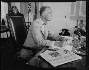 Gov. Christian Herter seated at desk with copy of Boston Herald with headline "Urges Herter Run with Ike"