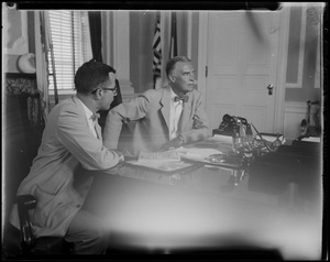 Gov. Christian Herter seated at desk with microphones, with man in glasses nearby