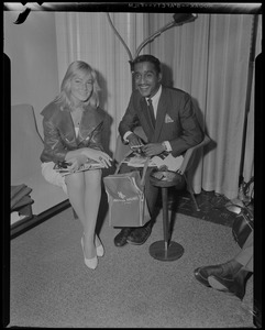 Sammy Davis, Jr., holding a cigarette and American Airlines bag, seated with May Britt