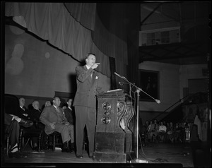 Billy Graham speaking at podium and pointing, with others seated on stage