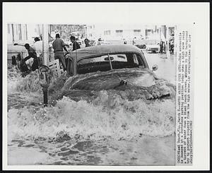 Waves Washes Over Auto--This automobile is swamped at a curbstone parking position today when a high wave sends a torrent of water down a Miami Beach street. Workmen in the background try to protect property from the high water.