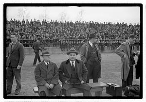 A group of men seated and standing, a crowd of spectators behind them