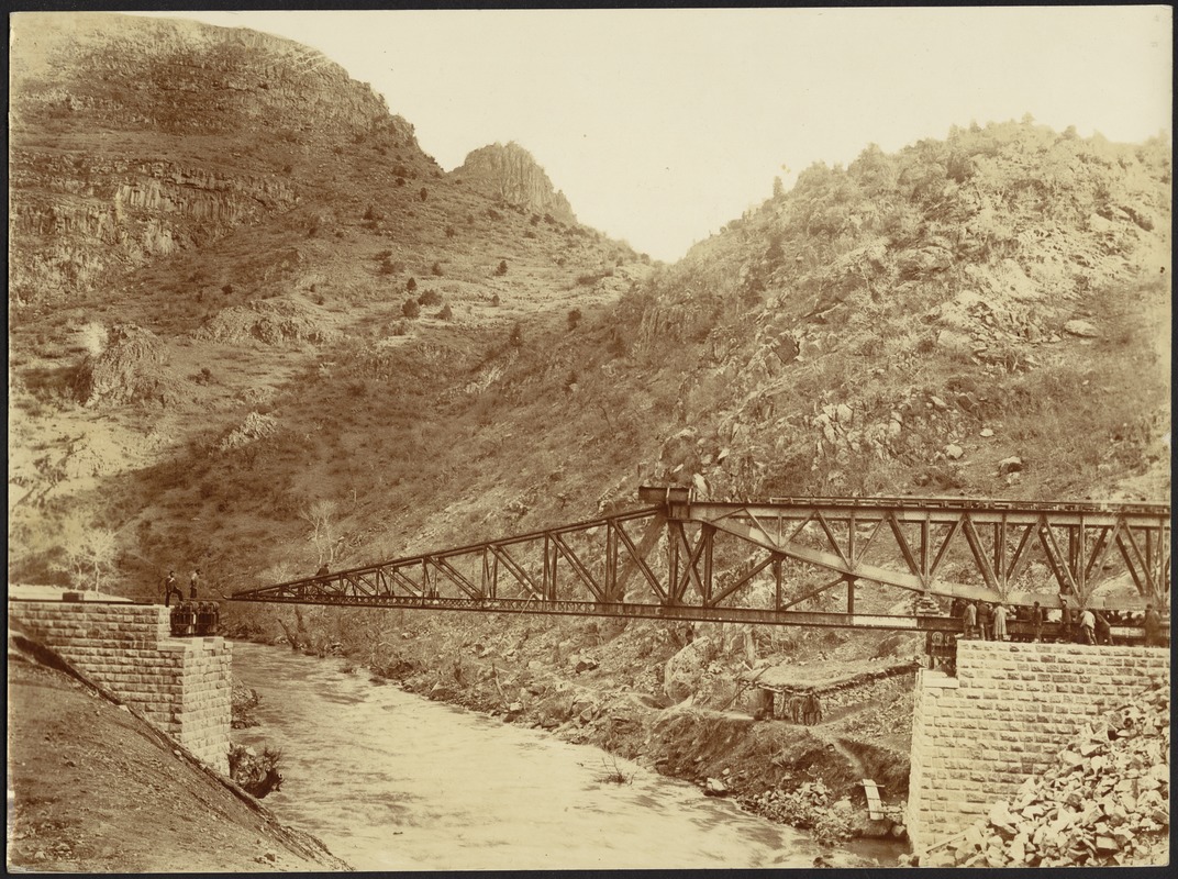 Construction of steel bridge over a river with men standing on stone anchorage