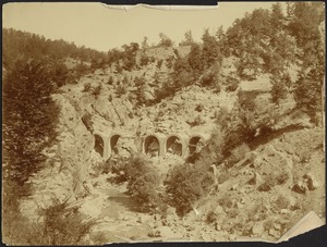 Construction of arched viaduct in river valley; people carrying bundles on left