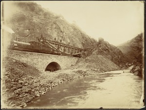 Section of steel bridge being transported by train across stone viaduct, men standing under arch