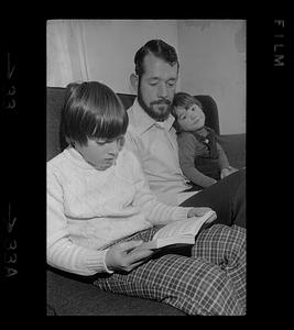 Boy reads on couch next to man holding toddler