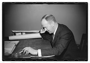 A man seated at a desk, blueprints in front of him