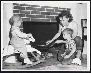 Indoors Cookout-With no electricity for stove, Mrs. Isabella Baltrusaites prepares dinner in frying pan over living room fireplace at home on Trafton road, Framingham, flooded by river water. Daughter Sandra, 17 months, on horse, and son Vincent, 3rd, look on.