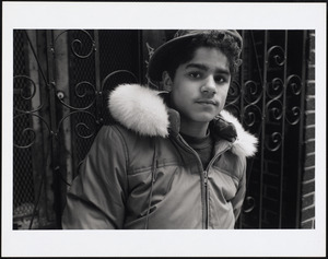 Young man wearing baseball cap stands in front of gate, probably Green Street, Jamaica Plain