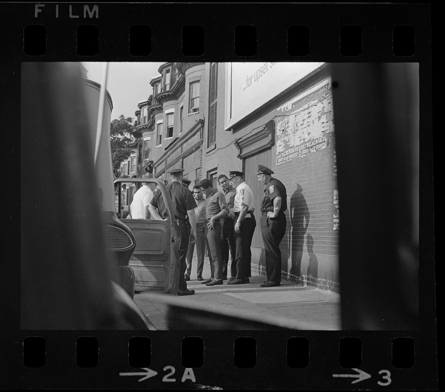 Police standing outside of a patrol car and speaking with a group of young Black men on the sidewalk, most likely after unrest broke out during student demonstrations