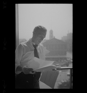 Mayor White standing by a window in his City Hall office overlooking Faneuil Hall and reviewing papers