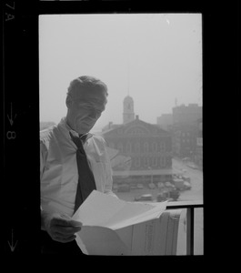Mayor White standing by a window in his City Hall office overlooking Faneuil Hall and reviewing papers