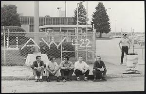 Picketers outside the only open entrance to Framingham’s General Motors assembly plant easing their feet. Union workers spelled out their Local number on the fence with discarded paper cups. A plant security officer stands apart from the group.