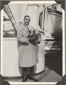 A man holding a Boston terrier with a lifeboat in the background