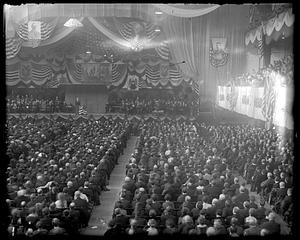 A large audience seated in front of a stage decorated with American flags