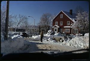 A person standing next to a dog in a snowdrift by a car parked in front of a house