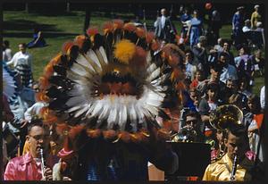 Back view of conductor wearing feather headdress facing musicians