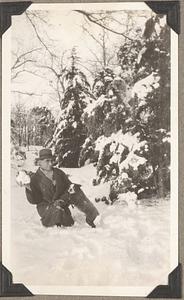 A man holding a snowball sits in the snow with a Boston terrier