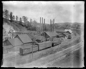 Train cars on railroad tracks next to building with smokestacks