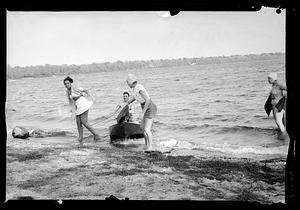 Three women walk out of the water while a man paddles his boat onto the beach