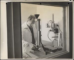 Interior view of text room through window showing technician testing surface drying rates of artists' oil paints with the sand testing machine