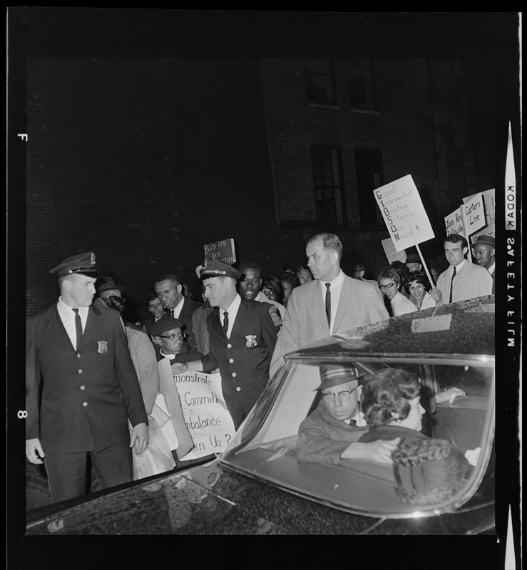 Reverend Vernon E. Carter watches car holding Sgt. John Hames and Louise Day Hicks during protest against school segregation outside Boston School Committee meeting