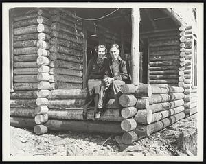 Modern Pioneers Launch Log Cabin Community. Spurred on by economic conditions, eight hardy families; reverting to the pioneer instincts of their fore fathers; have begun a log-cabin cooperative community on the edge of a wilderness that flanks the Daniel Webster Highway in this section. Among the contributing factors which led the twentieth century pioneers to strike out for themselves were high rents; cramped play room for children and lack of farming space. The cabins, built by the settlers themselves at a cost of less than $400; are constructed of pine; with clear maple and white birch flooring. All the timber supply is taken from the surrounding woods. The original cost of the land; about $60 an acre, and materials not available from natural sources constitute the outlay for what members of the community describe as "comfortable homes." Photo Shows: One of the couples of the log-cabin community... Mr. and Mrs. Burton Smith, seated on the porch railing of their nearly-completed little nest in the woods, where they will celebrate their first wedding anniversary in January. They expect to have everything in readiness before the first fall of snow, and next year they will cultivate the acre of land surrounding their cabin.