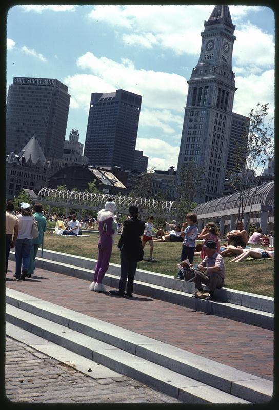 People sitting on grass, Christopher Columbus Park, Custom House Tower in background, a man dressed as a clown in foreground