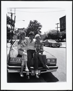 Two women and a man sit together on the hood of a car