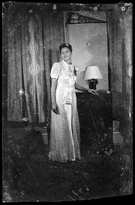 A woman in a floor length dress stands by a lamp
