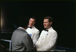 Man in gray jacket facing two men in white jackets and bow ties