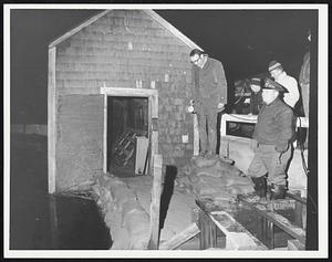 Men look out at water, one standing on sandbags