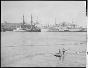 Two boys on homemade raft, Boston Harbor, Navy Yard behind them