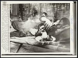 Problems Go Up in Smoke-A man contentedly smokes his opium pipe in corner of American run hospital at Ban Son, Laos, as small girl, just visible over man's elbow, plays in background. The man is a civilian, one of those evacuated from a hospital at Long Cheng shortly before an attack by North Vietnamese troops. Like thousands of Meo and Lao Tung tribesmen, he has joined a growing flood of refugees fleeing before [illegible]