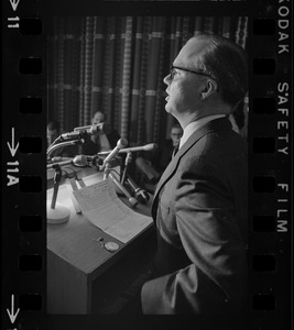 A man seen from behind, with his notes visible, speaking from a podium