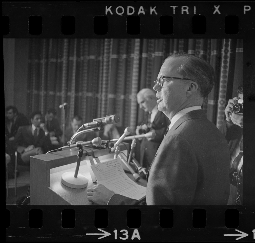 A man seen in profile, with his notes visible, speaking from a podium